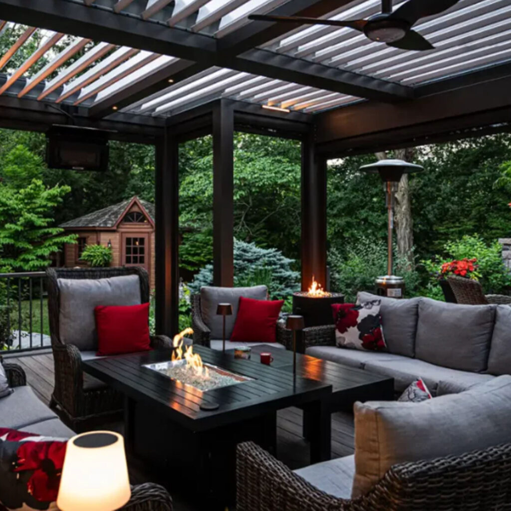 Cozy outdoor living space under a louvered pergola with wicker furniture, fire pit table, ambient lighting, and stone fireplace – backyard project by The Deck Store.