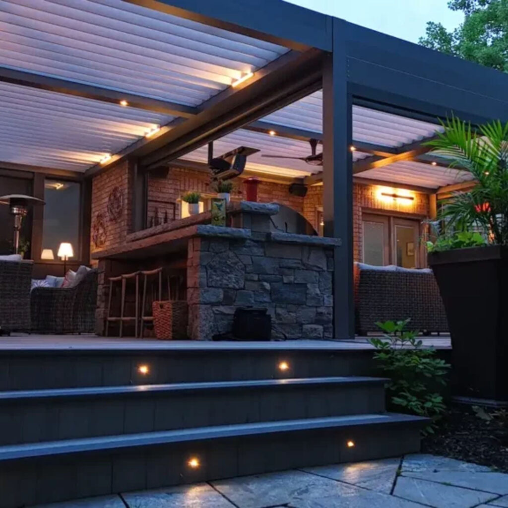Modern outdoor kitchen and lounge with stone bar, under a louvered pergola with ambient lighting and stair lights – backyard renovation by The Deck Store